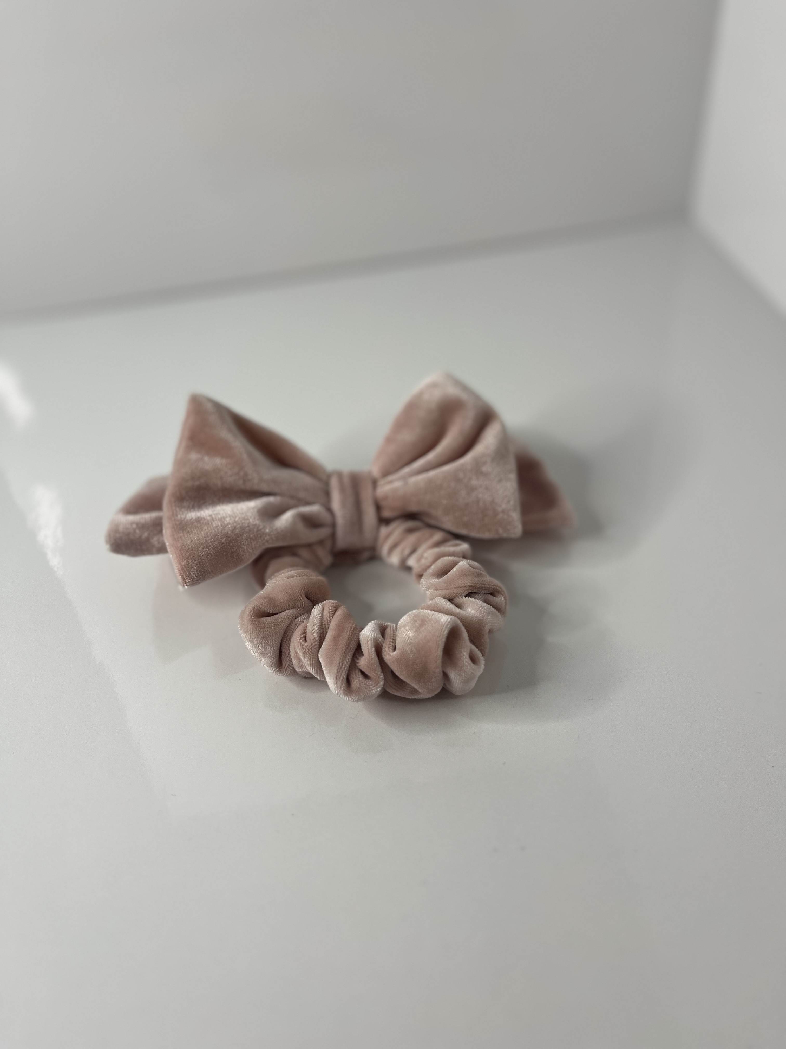 A pink velvet scrunchie with a bow rests on a flat surface, its torus shape accentuated by soft folds.