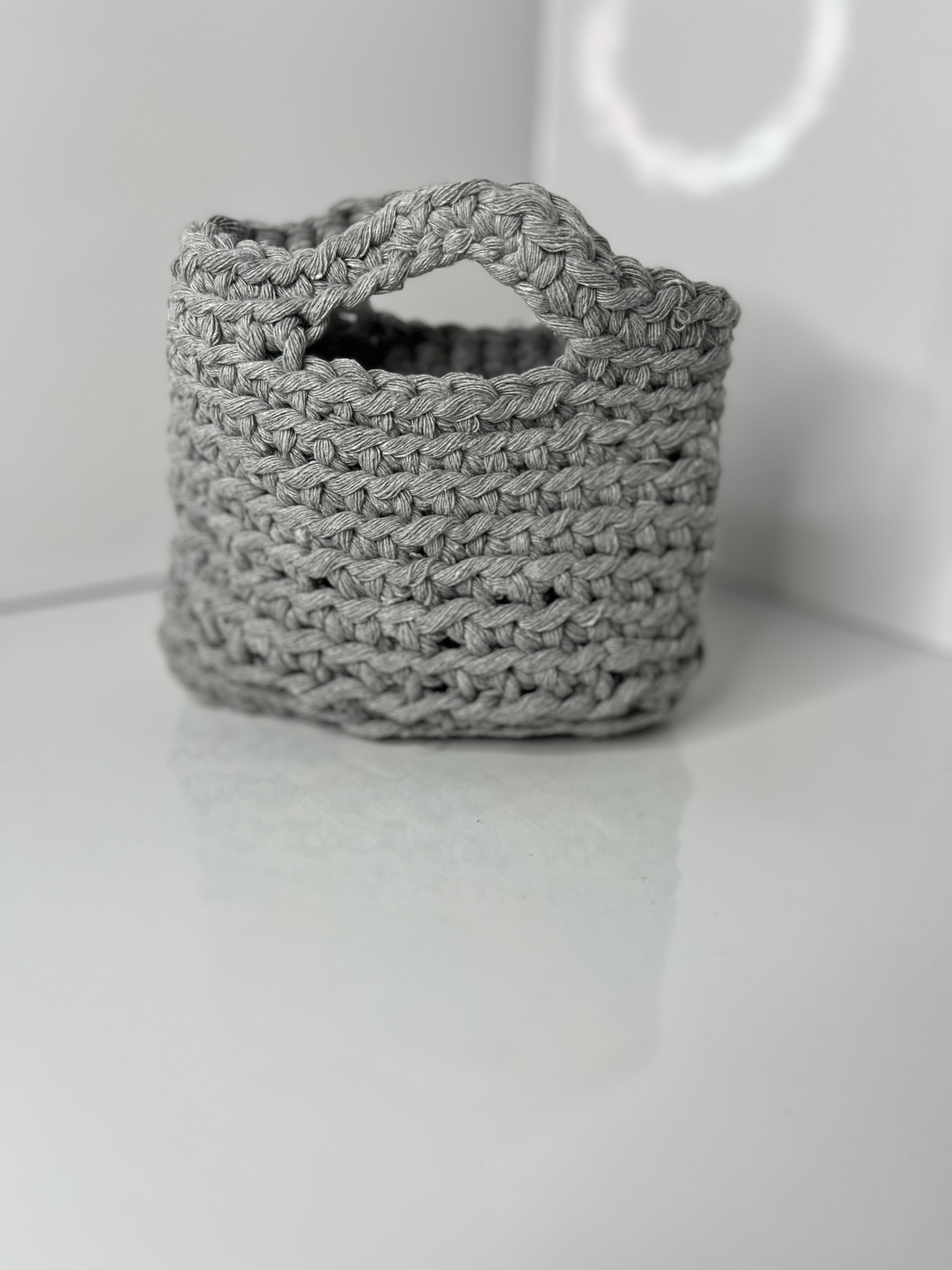 A gray hand-crocheted mini bag is photographed from an angled view, emphasizing the woven texture and handle opening.