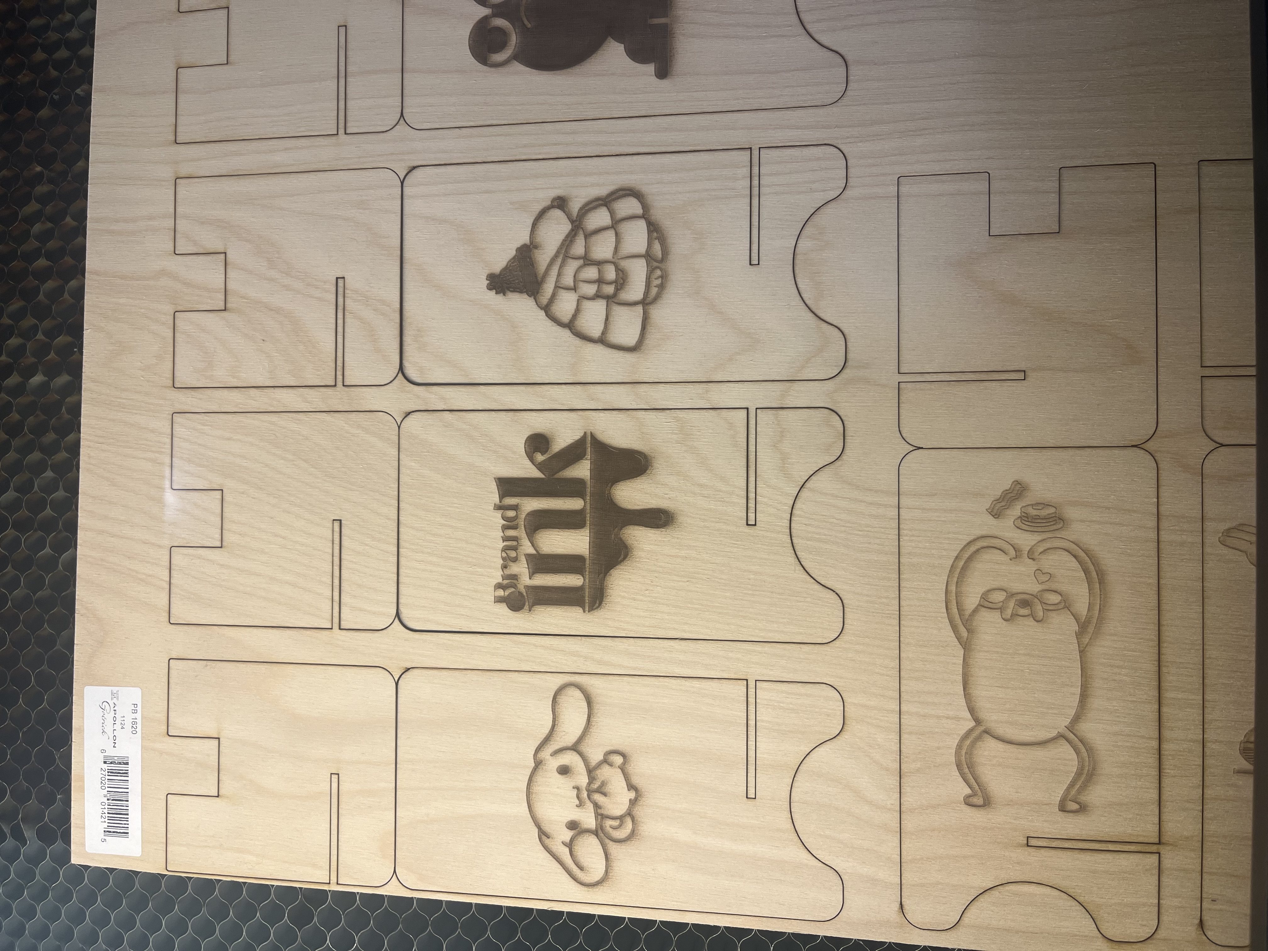 Four individual laser-cut plywood phone-stand components are displayed, including two structural base pieces and two engraved decorative panels featuring illustrated characters.